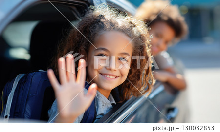 Smiling girl with backpack waving happily from car window on a sunny day 130032853