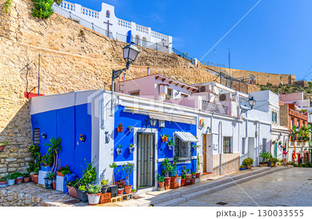 Alicante old town Santa Cruz with narrow street, typical white houses with flowers and plants and Ermita de la Santa Creu d'Alacant church in Alicante city historic centre, Valencian Community, Spain 130033555