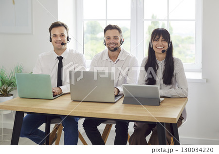 Smiling customer support agents with headsets and laptops sitting by window 130034220