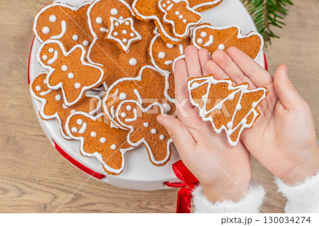 Hands holding Christmas tree shaped gingerbread cookie with icing over festive plate, top view 130034274