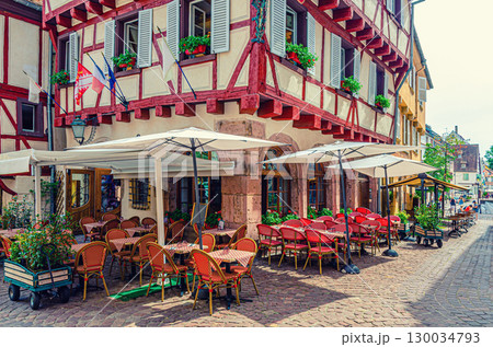 Street restaurant outdoor cafe tables, chairs and umbrellas, old colorful building half-timbered facade and shutters windows in old town Colmar city historic centre, Alsace Grand Est region, France Street restaurant outdoor cafe tables, chairs and umbrellas, old colorful building half-timbered facade and shutters windows in old town Colmar city historic centre, Alsace Grand Est region, France 130034793