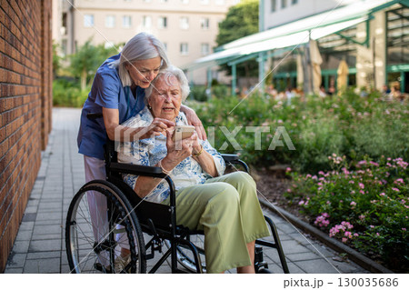 Senior patient in wheelchair showing picture in smartphone to caregiver. 130035686