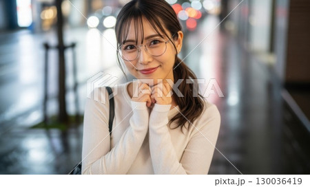 夜の街で両手を頬に当て、可愛らしく微笑むメガネ女子 130036419