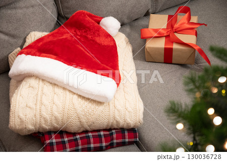 Christmas red Santa hat and knitted sweater, gift box with red ribbon on couch. Christmas tree and festive atmosphere. 130036728