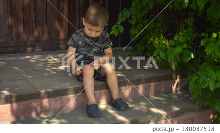the child touches a scab on his knee with his finger while sitting on the porch on a hot summer day the camera slowly moves closer he is wearing a t shirt and shorts 130037518