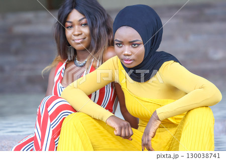 Two African women in colorful outfits enjoying a moment together outdoors in a relaxed setting 130038741