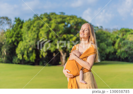Mother holding her little baby in a yellow sling in the park. Warm and loving family moment. Babywearing, parenting, and mother-child bonding concept Mother holding her little baby in a yellow sling in the park. Warm and loving family moment. Babywearing, parenting, and mother-child bonding concept 130038785
