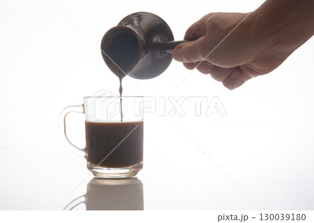 A hand pours rich coffee from a traditional ceramic pot into a clear glass cup A hand pours rich coffee from a traditional ceramic pot into a clear glass cup 130039180
