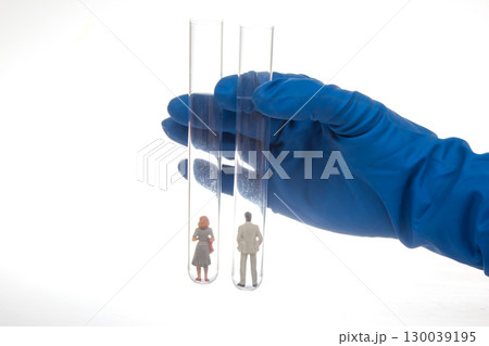 A gloved hand holds two test tubes containing miniature figurines of a man and woman 130039195