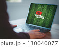 Person watching a live football match on a computer laptop screen with a green football field background. Concept of online sports streaming, digital entertainment, or live sports broadcast at home. 130040077