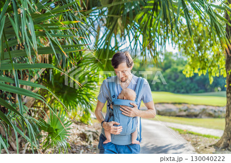 Young father walks in nature with his baby son nestled close in a parent-facing ergonomic baby carrier. Bonding, warmth, and modern parenting concept in a peaceful outdoor setting 130040222