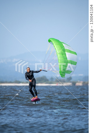 A middle-aged man in a wetsuit glides on a hydrofoil board with a green and white kite 130041104
