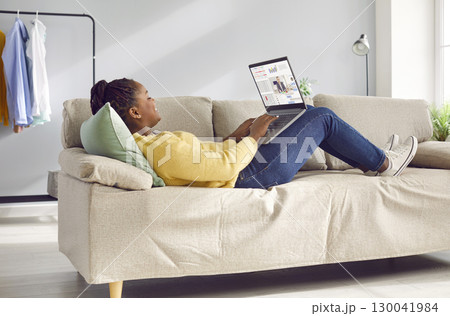 Happy young African American woman working remotely, lying on sofa and using laptop 130041984