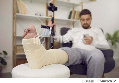 Man with bandaged leg using mobile phone while sitting in armchair Man with bandaged leg using mobile phone while sitting in armchair 130042012