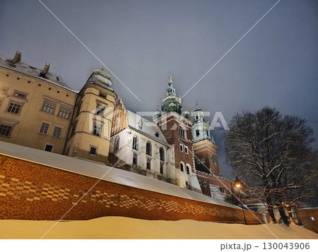 Snow covers the ground in front of an illuminated, historic building with multiple spired towers, a part of a Krakow castle 130043906