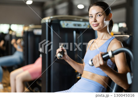 Positive young girl training on arm curl machine in gym Positive young girl training on arm curl machine in gym 130045016