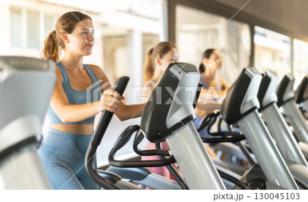 Positive young girl training on elliptical machine in gym Positive young girl training on elliptical machine in gym 130045103