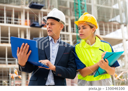 Man engineer standing on a construction site with a young woman worker monitors the construction plan on a laptop 130046100