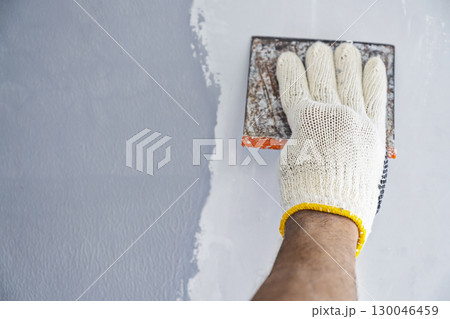 Construction worker in gloves using a scraper to fill cracks and surface imperfections on the wall during home renovation. Home renovation project Construction worker in gloves using a scraper to fill cracks and surface imperfections on the wall during home renovation. Home renovation project 130046459