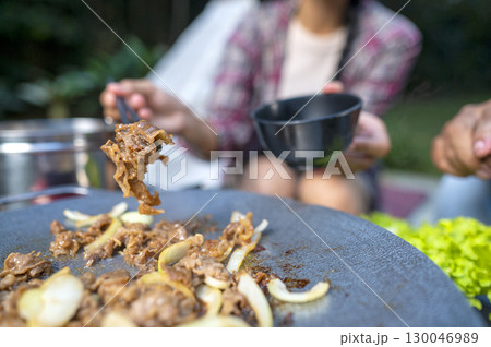 Closeup view of people with chopsticks cooking sliced marinated beef bulgogi on a round grill pan with onions. The scene of the lifestyle of enjoying food in a natural setting Closeup view of people with chopsticks cooking sliced marinated beef bulgogi on a round grill pan with onions. The scene of the lifestyle of enjoying food in a natural setting 130046989