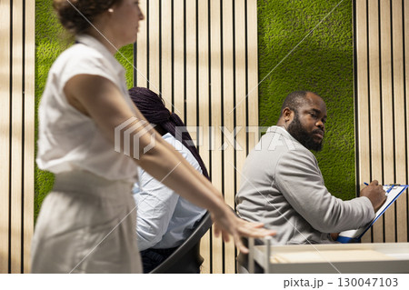 Businesswoman secretary walking with trolley cart and collecting CVs from people sitting in line on the hallway, corporate onboarding process. Candidates preparing for career opportunity. 130047103