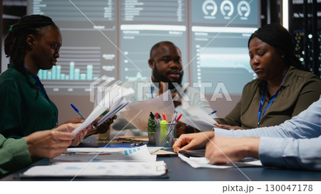 Close up of diverse specialists evaluating data numbers on archived files, exchanging reports and insights to establish clear enterprise objectives in strategy room late at night. Camera A. 130047178