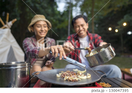 Indonesian southeast asian couple with tongs cooking sliced marinated beef bulgogi on a round grill pan with onions. The scene of the lifestyle of enjoying food in a natural setting 130047395