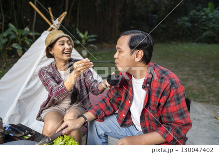 Indonesian southeast asian couple enjoying a meal with chopsticks. Smiles and enjoying togetherness while grilling and camping outdoors. The scene of the lifestyle of enjoying food in a natural Indonesian southeast asian couple enjoying a meal with chopsticks. Smiles and enjoying togetherness while grilling and camping outdoors. The scene of the lifestyle of enjoying food in a natural 130047402