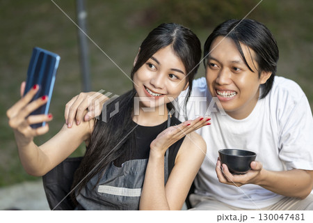 Indonesian southeast asian couple capturing a selfie using a cellphone. Woman holds the phone, with man beside her and holding a bowl. The scene of the lifestyle of enjoying food in a natural setting 130047481