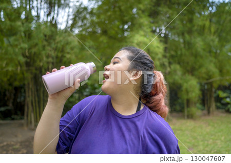Indonesian southeast asian overweight woman in a sporty outfit drinking water after practice running exercise in a public park outdoors. Concept of a Healthy active lifestyle and body inclusivity 130047607