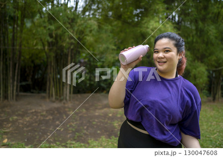 Indonesian southeast asian overweight woman in a sporty outfit drinking water after practice running exercise in a public park outdoors. Concept of a Healthy active lifestyle and body inclusivity 130047608