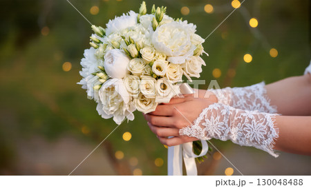 Bride holding elegant white wedding bouquet with roses and peonies, hands in lace gloves, outdoors with soft bokeh lights. Bride holding elegant white wedding bouquet with roses and peonies, hands in lace gloves, outdoors with soft bokeh lights. 130048488
