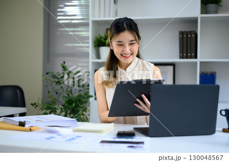 Smiling businesswoman reviewing documents while working with laptop in modern office Smiling businesswoman reviewing documents while working with laptop in modern office 130048567