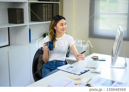Young Asian woman holding coffee mug while working at desk with computer in modern office Young Asian woman holding coffee mug while working at desk with computer in modern office 130048569