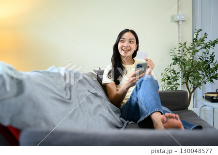 Relaxed woman using mobile phone and debit card to make online payment at home Relaxed woman using mobile phone and debit card to make online payment at home 130048577