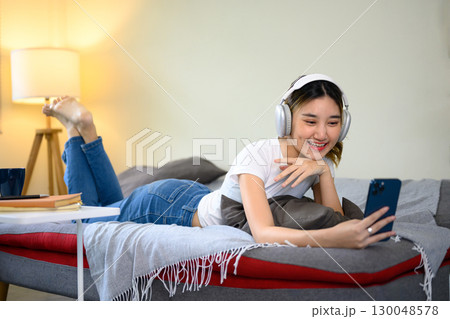 Relaxed Asian girl using phone and headphones for online chat at home 130048578