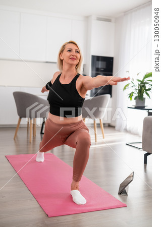 Middle aged blonde woman in sportswear standing in warrior pose on pink yoga mat in bright apartment, focusing on breathing and mindfulness 130050948