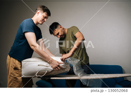 Two young men working together, using vacuum cleaner to pack bedding into vacuum storage bag placed on ironing board, efficiently organizing living space at home. Concept of household chores. Two young men working together, using vacuum cleaner to pack bedding into vacuum storage bag placed on ironing board, efficiently organizing living space at home. Concept of household chores. 130052068