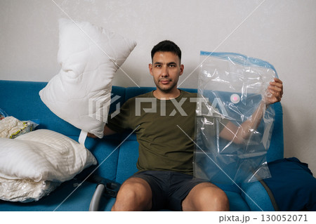 Portrait of young man packing white pillows and bedding into vacuum storage bags sitting on blue sofa in living room, getting ready for moving or tidying up. Concept of space saving storage. 130052071