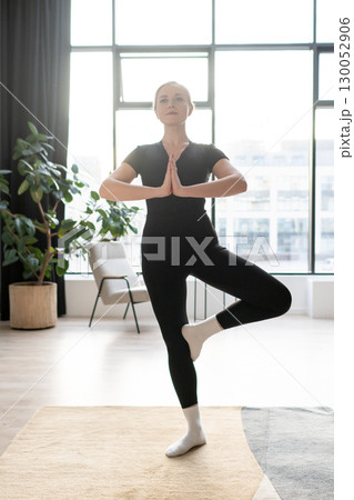 Woman in black sportswear practicing tree pose during yoga session in a bright room with large windows. 130052906