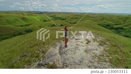 Young woman standing on top of a hill with open arms, embracing the beauty of rolling green hills and lush vegetation of Sumba Island under a cloudy sky 130053311