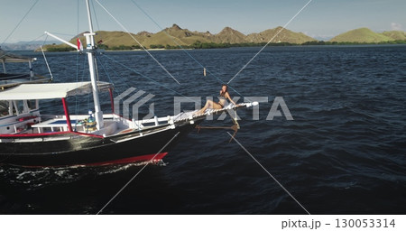 Aerial drone footage of a young tourist relaxing and sunbathing on the bowsprit of a yacht sailing in the beautiful Komodo National Park, Indonesia 130053314