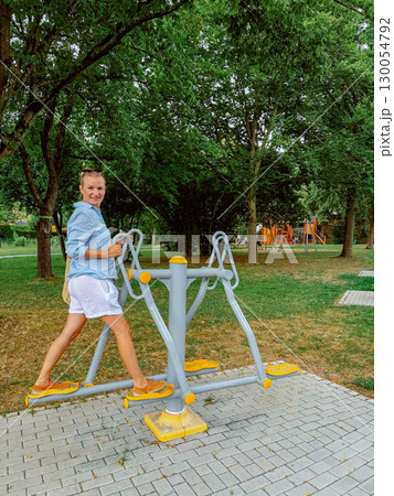Woman using outdoor fitness equipment in a park setting. Her expression is cheerful and focused on her workout. Surrounding greenery provides a pleasant environment for exercise. 130054792
