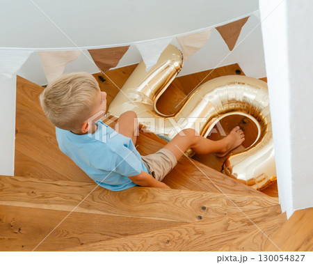 A young boy wearing a blue shirt and beige shorts sits on wooden stairs, holding a large gold-colored balloon in the shape of a number 6. Party decorations hang above. 130054827