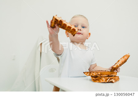 A happy baby joyfully enjoys a delicious waffle at the table, surrounded by a cheerful atmosphere that radiates happiness, showcasing precious moments of childhood and family togetherness 130056047