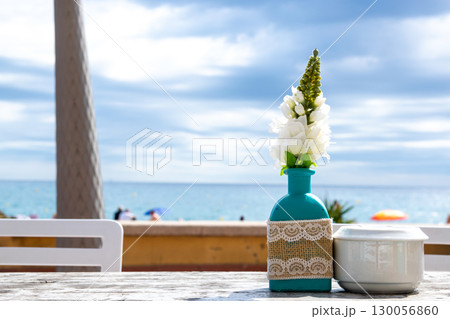 Warehouse on the open air with people. Chairs and tables outside on the summer sun weather. Vase with the flowers on the table for summer cafe with sea view in Spain. 130056860