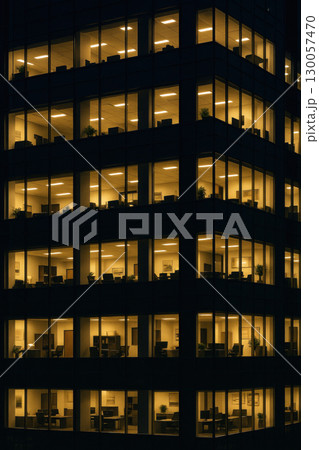 Office building at night with illuminated windows showing empty workspaces and desks in modern corporate environment 130057470