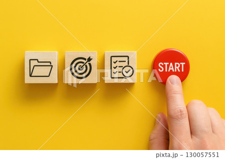 Finger pressing red start button beside wooden blocks with folder target and checklist icons project initiation concept on yellow background 130057551