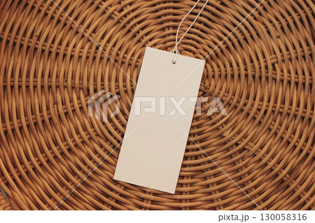 Close-up of blank white paper gift tag with string on brown woven wicker background. Minimal branding design. Price label, retail, product packaging. Fashion tag mockup. Copy space, flat lay, top view 130058316