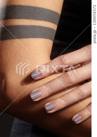 Close-up photo of a female hand, focus on gray manicure, neat short nails with gel polish. Hand resting on forearm with simple tattoo of two black stripes 130058372